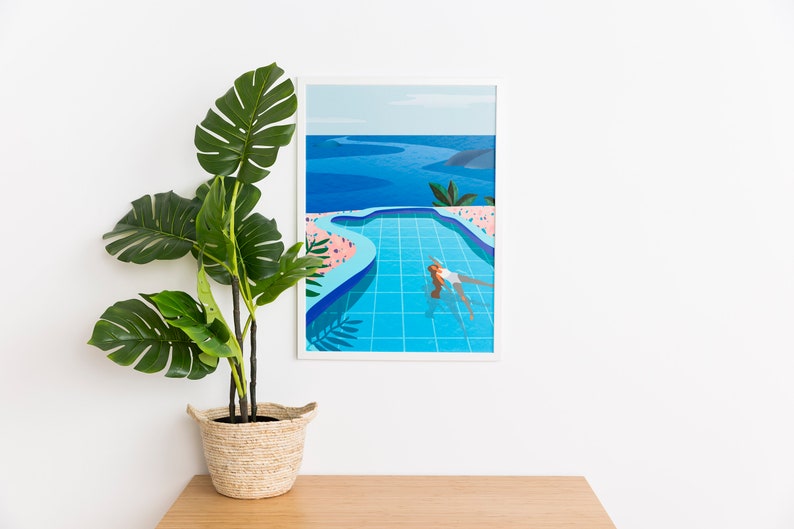 May include: A framed art print showing a person swimming in a turquoise pool with an ocean view. The artwork uses a minimalist style with blue, pink, and green colours. A large potted plant with green leaves is positioned to the left of the artwork.
