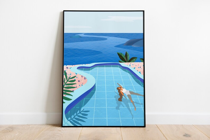 May include: A framed art print depicting a person in a blue swimming pool. The pool is set in a landscape of pink and blue hues, with a sea in the background. The artwork has a modern, minimalist aesthetic.