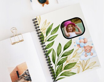 May include: A spiral bound notebook with a white background and a green and gold leaf design. The notebook has a photo of a young girl wearing a pink hat and a teddy bear holding balloons on a cloud.