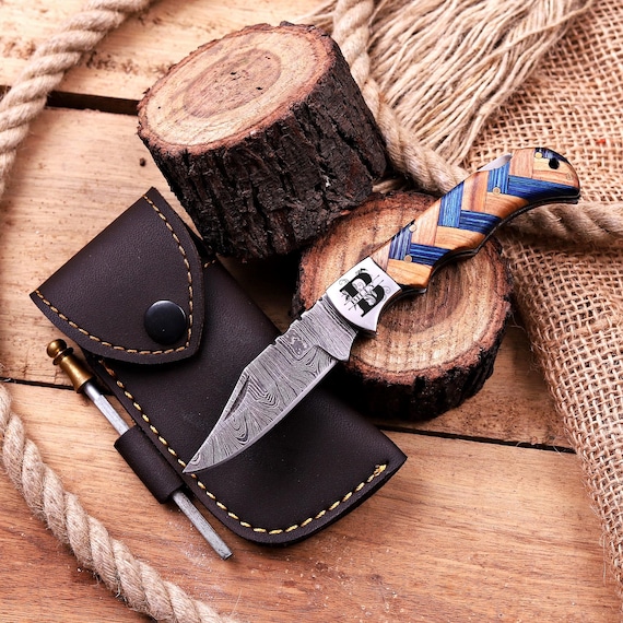 A hand-forged Damascus steel folding pocket knife with custom engraving — the swirling layered steel pattern alone makes it a showpiece, not just a tool.