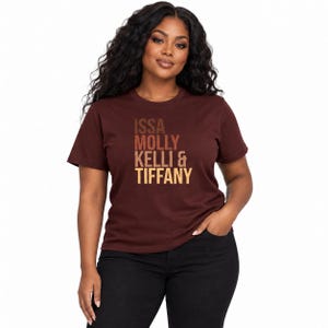 Op de afbeelding: Een donkerbruin T-shirt met korte mouwen en de tekst ISSA MOLLY KELLI & TIFFANY in een verticaal kleurverloop van bruin, beige en crème. Het T-shirt heeft een ronde hals en een standaard pasvorm, gedragen met een zwarte broek.