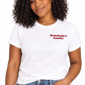 May include: A white short-sleeved t-shirt featuring the text Somebody's Auntie. printed in a dark red font on the upper left chest area. The garment has a classic crew neckline and a standard fit, paired with blue denim jeans with a metal button.
