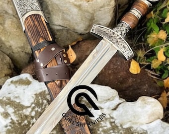 May include: A medieval-style sword with a silver blade and ornate handle, resting on a rock. The sword has a wooden handle and sheath with decorative metal accents. The sheath is brown with a leather strap.
