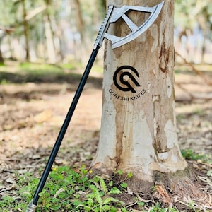 May include: A silver and black axe with a long handle is propped against a tree. The axe head has a unique design with a curved blade and geometric cutouts. The handle is black, and the axe is labeled with the text "QURESHI KNIFES."