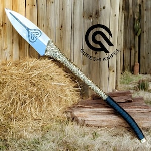 May include: A decorative spear with a silver blade and ornate handle. The handle features a detailed gold and silver pattern, transitioning to a dark blue wrapped grip. The spear is displayed with a hay bale and wooden backdrop. The logo "Qureshi Knives" is visible.