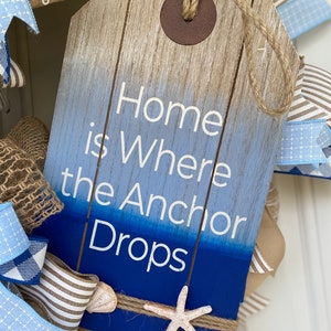 Close up of the wood wreath sign with the inscription Home is Where the Anchor Drops. It is shaped like an oversized gift tag with a wood plank background dipped in ombré blue paint. Includes several small shells and jute rope trim.