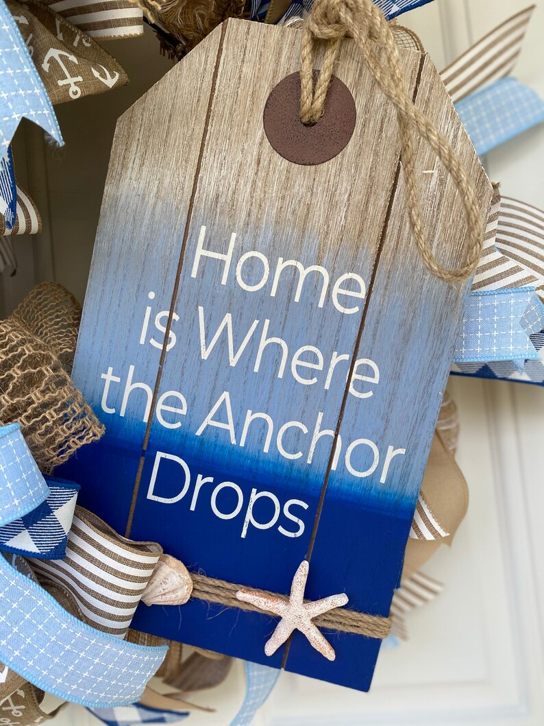Close up of the wood wreath sign with the inscription Home is Where the Anchor Drops. It is shaped like an oversized gift tag with a wood plank background dipped in ombré blue paint. Includes several small shells and jute rope trim.