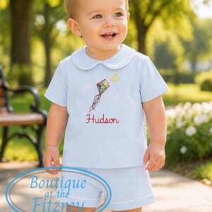 May include: A light blue short sleeve shirt and shorts set with white trim. The shirt features a kite and sun design with the name "Hudson" embroidered in red. The outfit is for a child.