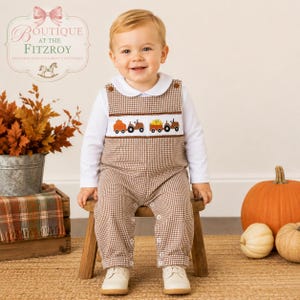 May include: A brown and white gingham overall with a white long-sleeve shirt. The overall features a smocked chest panel with embroidered orange pumpkins and brown tractors. The garment has a white Peter Pan collar and white buttons at the shoulders.