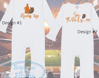 May include: Two white baby rompers with long sleeves and ruffle cuffs. Design #1 features an orange illustration of a bottle and football with "Rocky Top" text. Design #2 has "Go Vols" text and a football illustration.