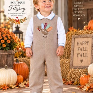 May include: A tan and white gingham overall with a turkey applique on the chest, featuring a football-shaped body and colorful tail feathers. The overall is worn over a white long-sleeve shirt with a light blue collar and cuff trim.