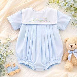May include: A light blue short-sleeve romper featuring a white sailor-style collar with an embroidered train motif. The garment has vertical pleats down the front bodice and elasticized leg openings, providing a structured yet soft appearance.