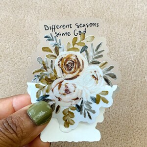 May include: A white sticker with a watercolor illustration of a bouquet of white and brown roses with green leaves. The sticker has the text "Different seasons same God" on it.