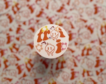 May include: A round sticker with a white background and orange text that says "tri" with a dripping smiley face. The sticker is surrounded by small flowers and smiley faces.