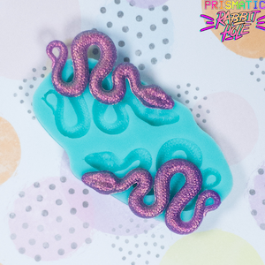 Puede incluir: Un molde de silicona para hacer dijes de resina en forma de serpiente. El molde es azul turquesa y presenta cuatro formas de serpiente. Dos serpientes de purpurina morada están sentadas encima del molde.