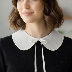 May include: A white Peter Pan collar with a delicate bow tie, set against a black top. The collar has a rounded shape and is made of a textured fabric. The bow tie hangs down from the center of the collar.