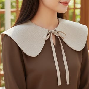 May include: A beige linen-textured collar with a rounded shape and a tie closure. The collar is worn over a brown long-sleeved top. The collar adds a vintage touch to the outfit.