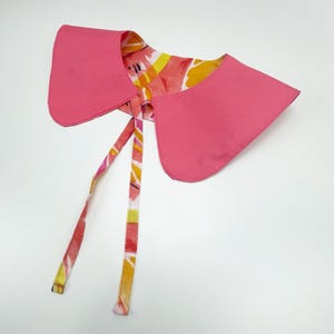 May include: A pink, detachable collar with a floral pattern on the reverse side. The collar has a rounded shape and long, patterned ties for securing. The floral pattern includes shades of red, yellow, and orange.