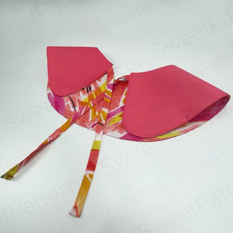 May include: A pink fabric collar with a rounded shape and tie closures. The collar's exterior is a solid pink, while the interior features a colorful abstract pattern in shades of pink, yellow, and orange. The ties are patterned.