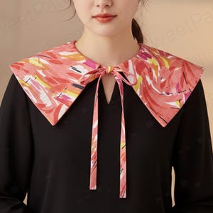 May include: A black top with a colorful, patterned collar. The collar features a vibrant design with pink, yellow, and white brushstrokes. A matching tie is knotted at the neckline, adding a decorative touch to the garment.