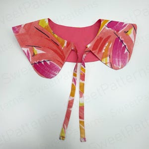 May include: A floral print collar with a pink backing and long ties. The collar features a vibrant pattern of pink, orange, and yellow brushstrokes. The ties are patterned with the same colors. The collar is designed to be worn around the neck.