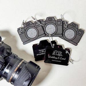 Puede incluir: Una colección de etiquetas grises con forma de cámara con centros de purpurina plateada y clips metálicos. Dos etiquetas negras muestran información de contacto y las palabras "Wedding & Event Photography" con un gráfico de cámara. Se ve una cámara Nikon con una lente Tamron.