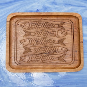 May include: A rectangular wooden serving board with a carved design of five fish. The board has a raised edge and a natural wood grain finish. The fish are detailed with scales and fins.