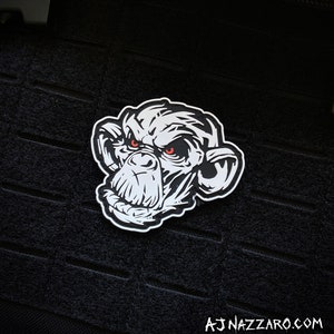 May include: A white and black patch featuring a detailed illustration of a chimpanzee's face with red eyes. The patch is set against a black background. The text "AJ NAZZARO.COM" is at the bottom.