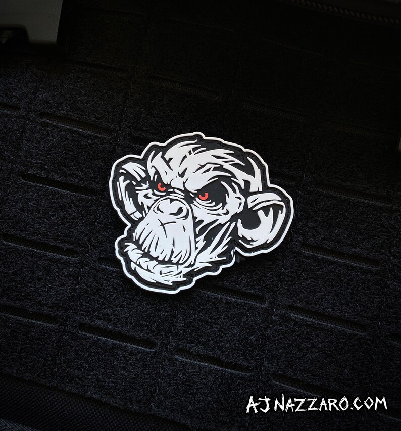 May include: A white and black patch featuring a detailed illustration of a chimpanzee's face with red eyes. The patch is set against a black background. The text "AJ NAZZARO.COM" is at the bottom.