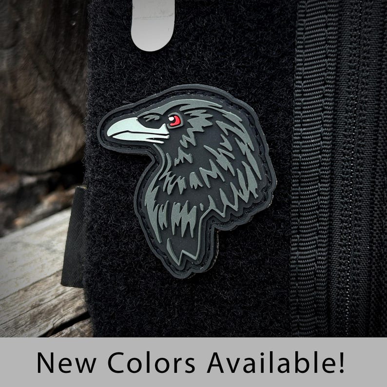 May include: Black and gray embroidered raven patch with red eyes. The patch is attached to a black fabric background.