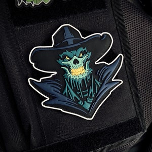 Pode incluir: Um remendo bordado preto e verde de uma caveira usando um chapéu de caubói. A caveira tem uma substância verde que pinga e um sorriso cheio de dentes. O remendo está preso a uma mochila tática preta.