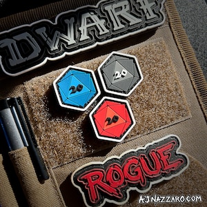 May include: Three 20-sided dice patches in red, blue and grey with white outlines. The patches are attached to a brown tactical vest with the words "DWARF" and "ROGUE" in white lettering.