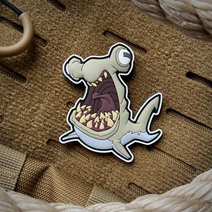 May include: A rubberized patch featuring a cartoon hammerhead shark with a wide-open mouth, sharp yellow teeth, and a dark purple interior. The shark is light tan with a white underbelly and a black outline. The text AJNAZZARO.COM is visible.