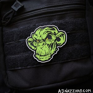 May include: A green and black patch featuring a stylized ape face with yellow eyes. The patch is affixed to a black tactical bag with velcro straps and a zipper. The text "AJ NAZZARO.COM" is visible at the bottom.