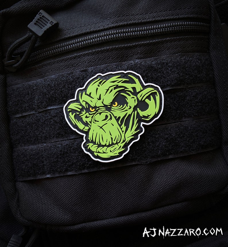 May include: A green and black patch featuring a stylized ape face with yellow eyes. The patch is affixed to a black tactical bag with velcro straps and a zipper. The text "AJ NAZZARO.COM" is visible at the bottom.