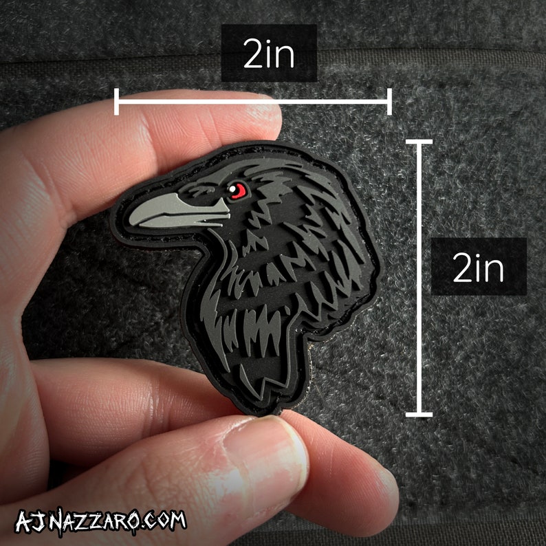 May include: A black and gray 2-inch by 2-inch embroidered patch of a raven with red eyes. The raven is facing left and has a detailed design.