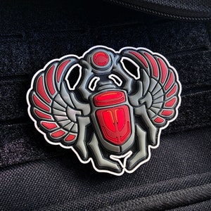 May include: A silver and red embroidered patch featuring a scarab beetle with wings. The beetle is outlined in white and has a red body with a white circle on its head.