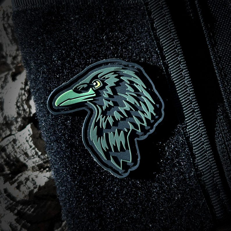 May include: A black and green embroidered patch of a raven's head with a green beak and eyes. The patch is attached to a black fabric background.
