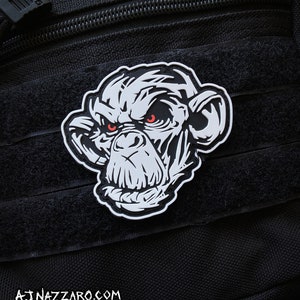 May include: A black and white patch featuring a stylized chimpanzee face with red eyes. The patch is set against a black background with a textured surface. The website address AJNAZZARO.COM is printed at the bottom.