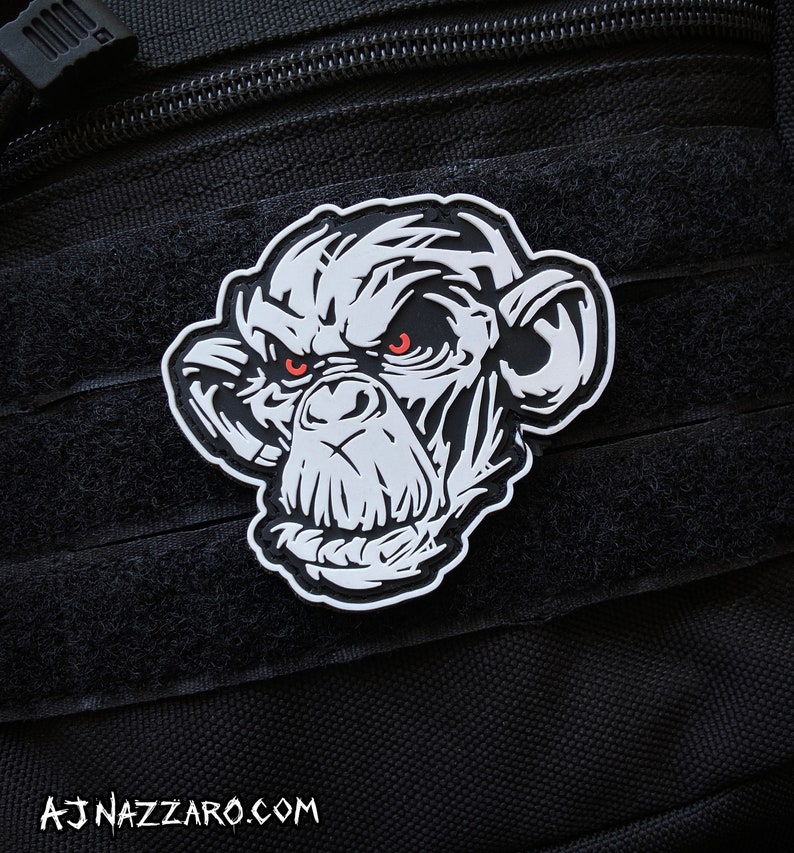 May include: A black and white patch featuring a stylized chimpanzee face with red eyes. The patch is set against a black background with a textured surface. The website address AJNAZZARO.COM is printed at the bottom.