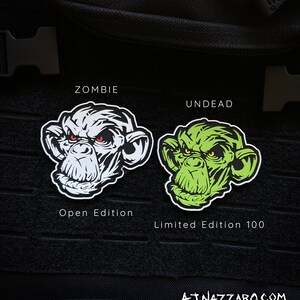 May include: Two patches featuring a stylized ape face. One is white with red eyes, labeled "ZOMBIE" and "Open Edition." The other is green with yellow eyes, labeled "UNDEAD" and "Limited Edition 100." The artist's signature is at the bottom.