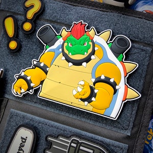 May include: A yellow, green and blue cartoon character with a red mohawk and spiky shell. The character is wearing a black spiked bracelet and has a menacing expression. The character is a patch with a white background.