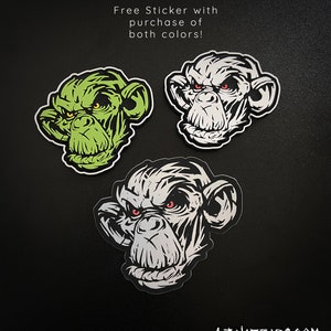 May include: Three stickers featuring a stylized monkey face with an angry expression. One is green, one is white, and one is gray. The stickers have red eyes and are on a black background. Text reads "Free Sticker with purchase of both colors!"