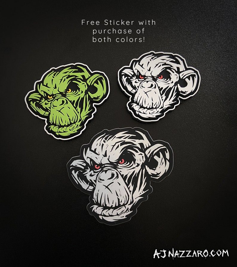 May include: Three stickers featuring a stylized monkey face with an angry expression. One is green, one is white, and one is gray. The stickers have red eyes and are on a black background. Text reads "Free Sticker with purchase of both colors!"