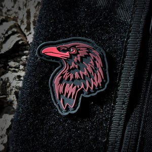 May include: A black and red embroidered patch of a raven's head. The patch is attached to a black tactical vest.