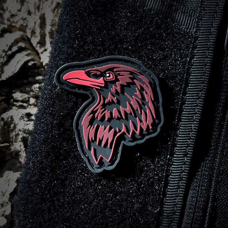 May include: A black and red embroidered patch of a raven's head. The patch is attached to a black tactical vest.