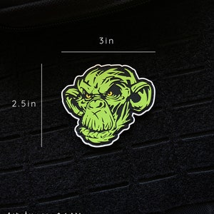 May include: A green monkey face sticker with yellow eyes and a white border. The sticker is 3 inches wide and 2.5 inches tall. The sticker is on a black textured surface. The website AJNAZZARO.COM is printed at the bottom.