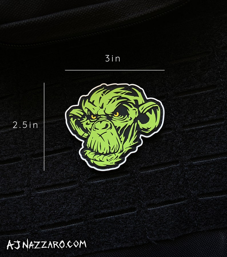 May include: A green monkey face sticker with yellow eyes and a white border. The sticker is 3 inches wide and 2.5 inches tall. The sticker is on a black textured surface. The website AJNAZZARO.COM is printed at the bottom.