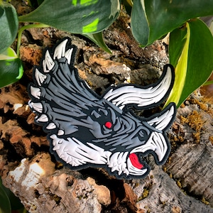 May include: A black and white embroidered patch of a triceratops dinosaur head with red eyes and a red mouth. The patch is on a brown wooden surface.