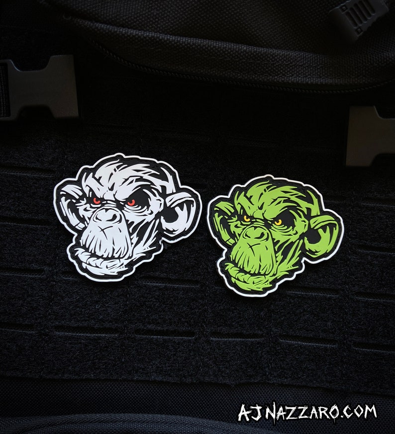 May include: Two monkey face patches. One is white with black details and red eyes, the other is green with black details and yellow eyes. The patches are on a black background. The text "AJNAZZARO.COM" is at the bottom.